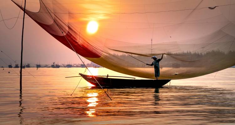 Bei Sonnenuntergang balanciert ein einsamer Fischer auf einem kleinen Holzboot und wirft ein großes Netz über schimmernde goldene Gewässer.