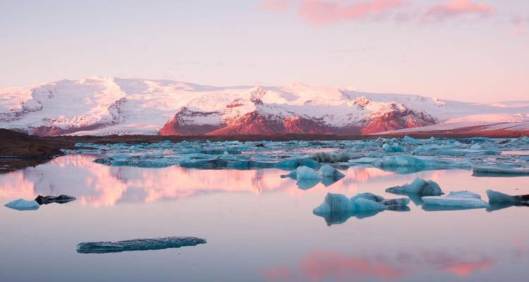 Los icebergs flotan en una laguna tranquila reflejando montañas de color rosa pastel al atardecer.