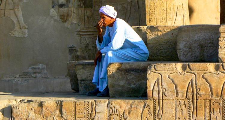 Un hombre con una ropa tradicional se sienta pensativo sobre bloques de piedra tallada de un templo antiguo.