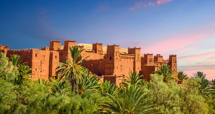 La forteresse en adobe d'Aït Benhaddou encadrée par des palmeraies luxuriantes sous un ciel rose en fin d'après-midi
