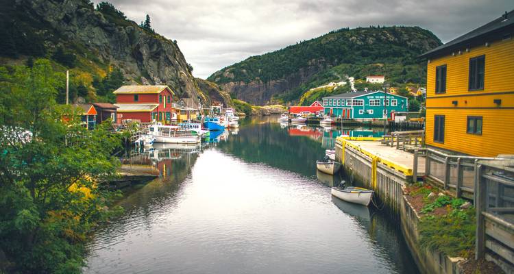 Narrow inlet lined with colourful wooden houses and fishing boats reflected in still water under overcast sky.