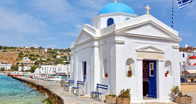 Small white seaside chapel with a blue dome and Greek flag beside clear waters