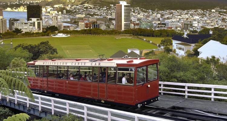De Red Wellington kabelwagen stijgt op boven de stadsgezicht en cricketveld met de haven in de verte.