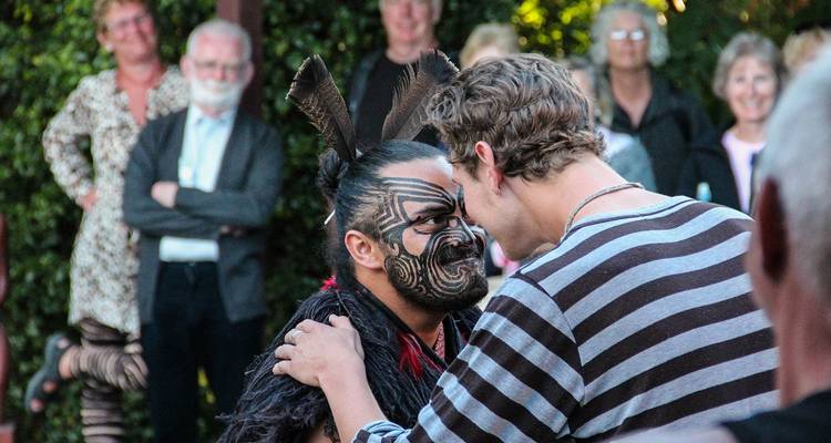 Maori warrior performs hongi greeting with a visitor while onlookers watch.