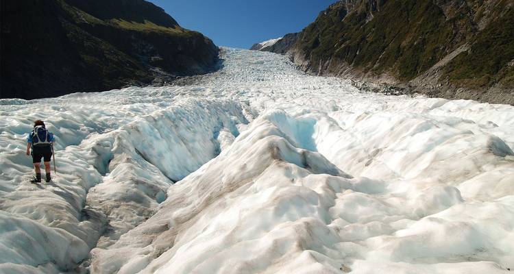 Hiker stands on the surface of a vast white and blue glacier surrounded by rugged peaks.