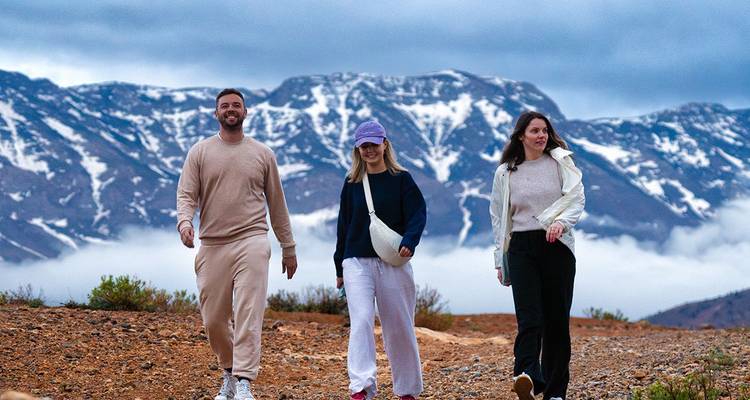 Drie reizigers die langs een rotsachtig plateau lopen met met sneeuw bestoven bergen op de achtergrond