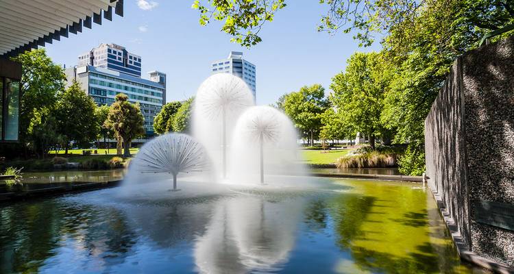Fontaine moderne avec trois jets en forme de pissenlit dans un bassin réfléchissant entouré de verdure luxuriante du parc urbain et de tours de bureaux.