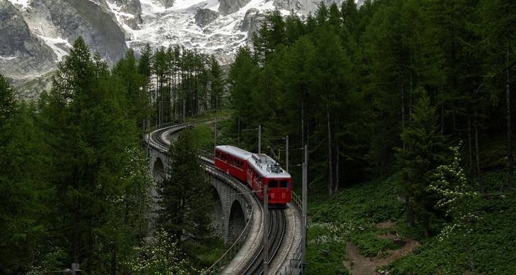 Un train de montagne rouge serpente à travers un viaduc élevé dans une forêt de conifères dense sous des pics enneigés escarpés.