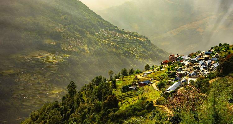Morning light spills over terraced green valleys and a small Nepalese hillside hamlet surrounded by lush slopes.
