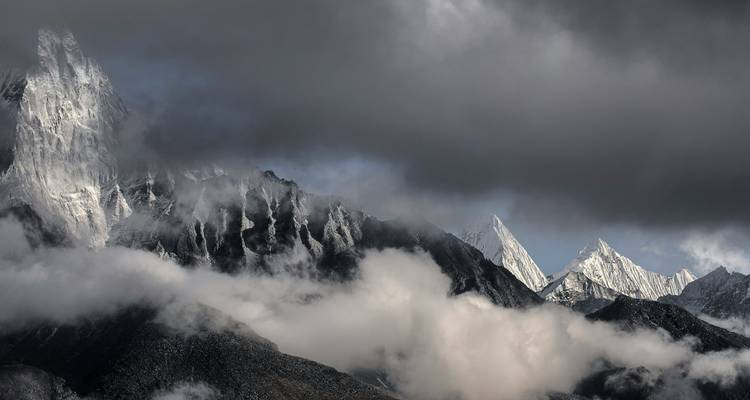 Des pics dramatiques et sombres de l'Himalaya enveloppés de nuages épais avec des rayons de lumière frappant les sommets déchiquetés.