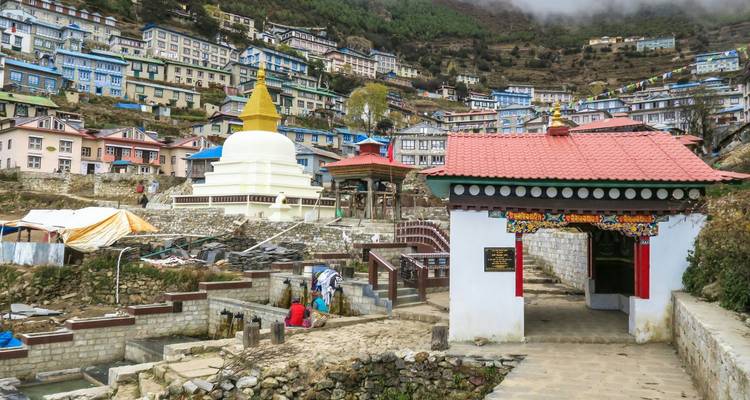 Ville de montagne construite sur une pente avec des maisons colorées, une stupa blanche et du brouillard roulant sur les crêtes.