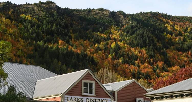 Rangée d'arbres aux couleurs automnales derrière des bâtiments rustiques en bois portant l'étiquette « LAKES DISTRICT » au pied d'une colline boisée.