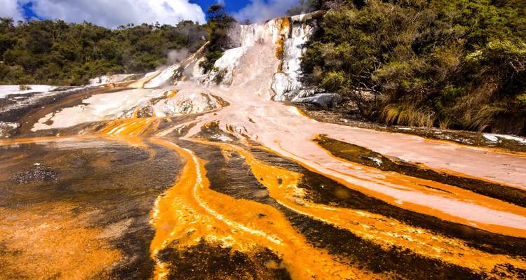 Leuchtend orange Mineralströme, die an geothermischen Hängen inmitten von Buschwerk herabfließen.