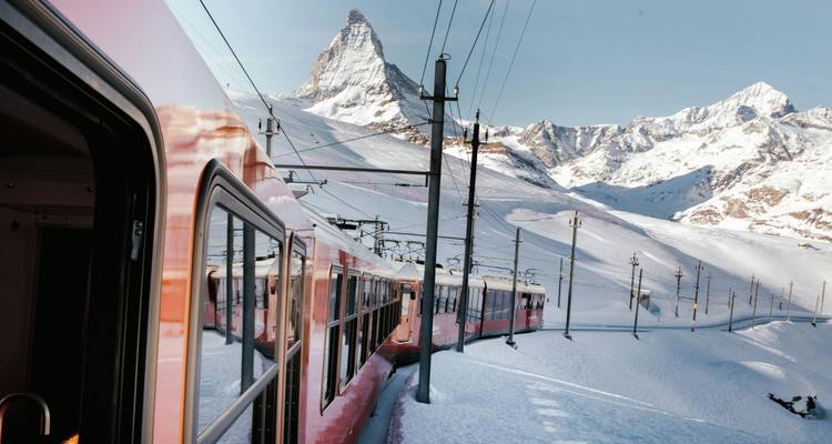 Red mountain train snakes through snowy alpine pass beneath a distinctive horn-shaped peak