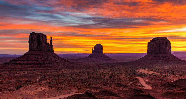 Tres icónicas formaciones rocosas del Valle del Monumento recortadas contra un cielo de amanecer naranja fuego y púrpura