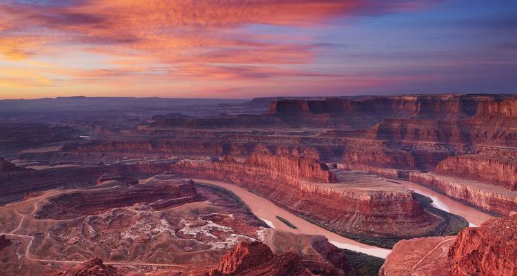 Un vasto cañón de roca roja esculpido por un río sinuoso brilla bajo una puesta de sol vibrante en tonos rosa y naranja