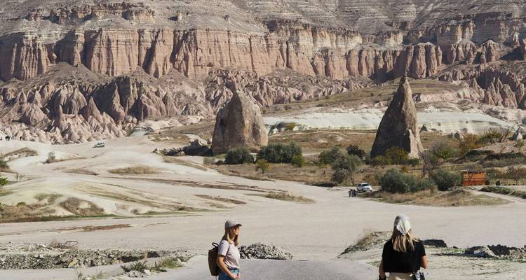 Two young women walk across a dramatic rocky valley with distinctive cone-shaped fairy chimneys and banded cliffs in the background.