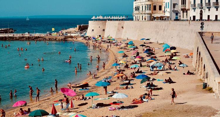 Busy sandy beach lined with colorful umbrellas and sunbathers beside calm turquoise water and an old sea wall.