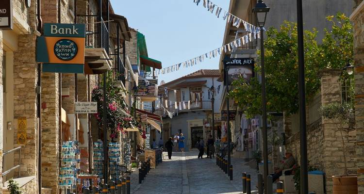 Charming stone-paved village street lined with shops, bunting, and blooming flowers under bright daylight.