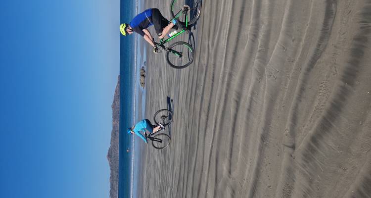 Pareja de ciclistas pedaleando sobre la arena mojada y ondulada de una playa ancha y vacía bajo un cielo azul despejado