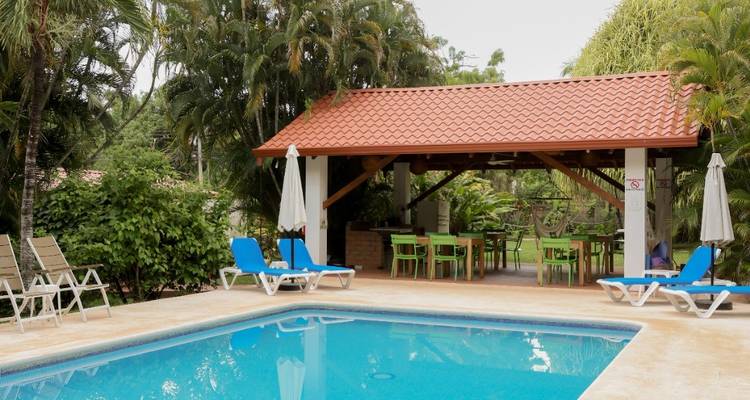 Pequeña piscina de hotel tropical con tumbonas y un pabellón comedor al aire libre con techo de tejas.