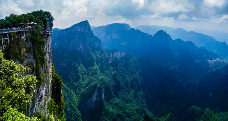 Cliff-edge walkway lined with tourists overlooking vast layers of green karst mountains.