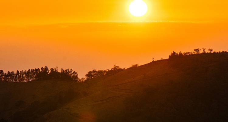 Brilliant orange sun sets over rolling hills, casting silhouettes and warm glow across the sky.