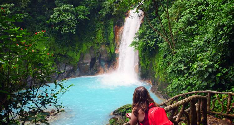 Traveler with red backpack admires the vibrant blue Rio Celeste waterfall in dense rainforest.
