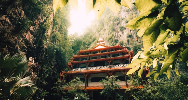 Templo tradicional enmarcado por exuberante vegetación y acantilados de piedra caliza que se alzan hacia el cielo, bañado por la suave luz del sol.