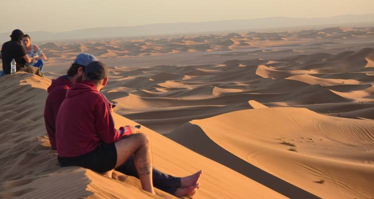 Travelers sit atop a high dune watching endless rolling sand seas at sunset in Erg Chigaga.
