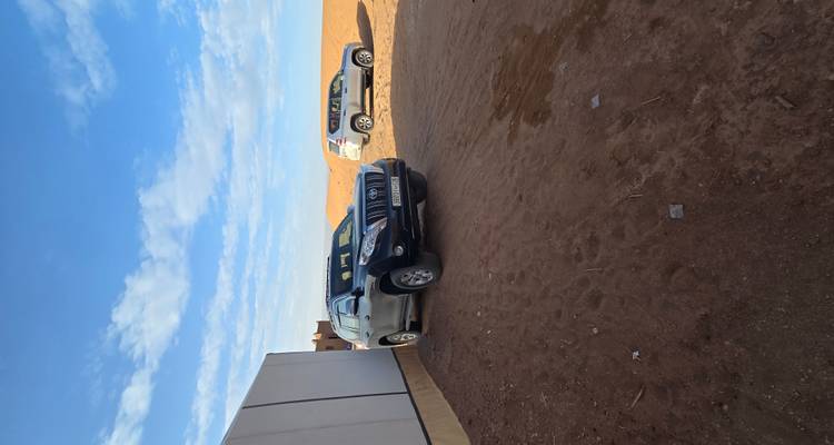 Two SUVs parked beside a desert camp wall under a bright blue sky and golden dune.