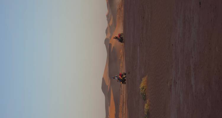 Two quad bikes ride across a flat desert plain with distant dunes at golden hour.