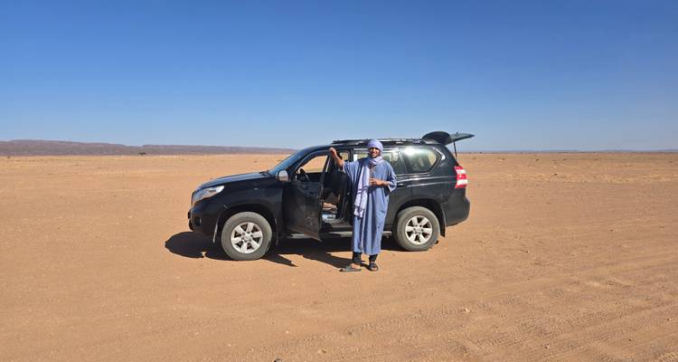 Local guide in traditional attire stands beside a 4x4 vehicle in an open desert plain.