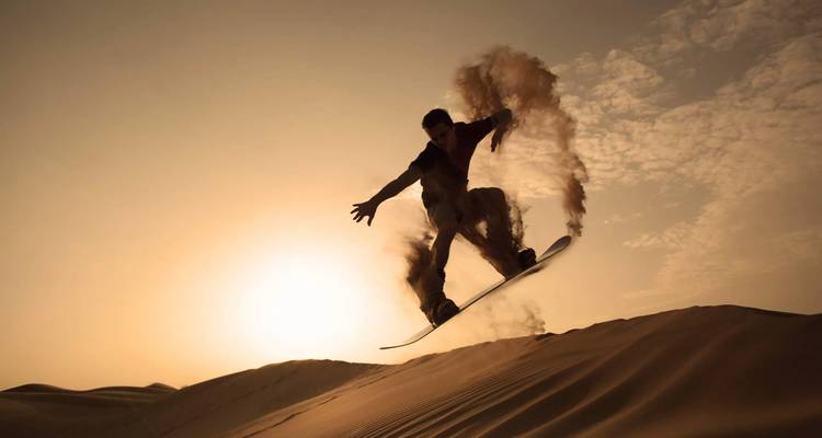 Silhouette d'un planchiste de sable en vol au-dessus d'une dune au coucher du soleil avec du sable traçant un arc