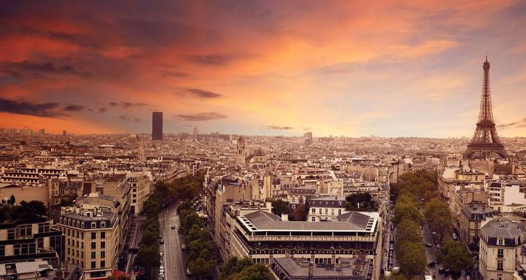 Panorámica amplia de París al atardecer con nubes dramáticas y la Torre Eiffel recortada contra el horizonte de los tejados de la ciudad.