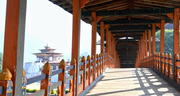 Pasarela cubierta de madera con pilares naranjas repetitivos que lleva hacia una dzong visible en la distancia brumosa.