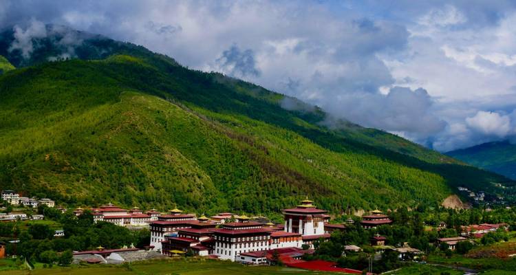 Valle amplio cubierto de bosque verde con un impresionante complejo de dzong en su base bajo nubes dramáticas.