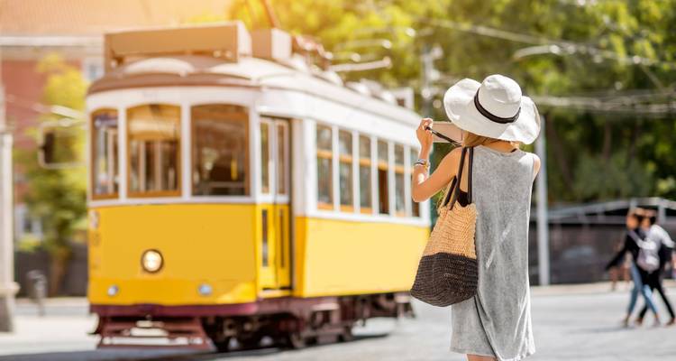 Woman in summer hat photographing a passing yellow tram on a sunny Lisbon street.