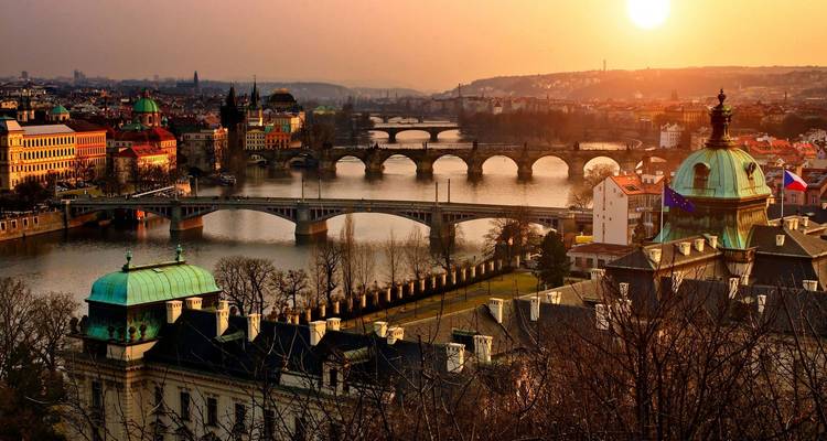 Coucher de soleil spectaculaire sur Prague avec plusieurs ponts traversant la rivière Vltava et des dômes historiques dans une lumière dorée chaleureuse.