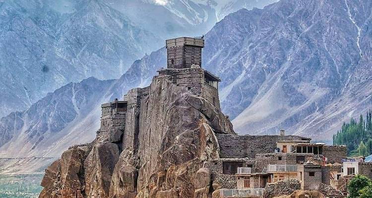Fortaleza de piedra encaramada en un afloramiento rocoso contra los dramáticos picos del Himalaya con casas de pueblos dispersas.