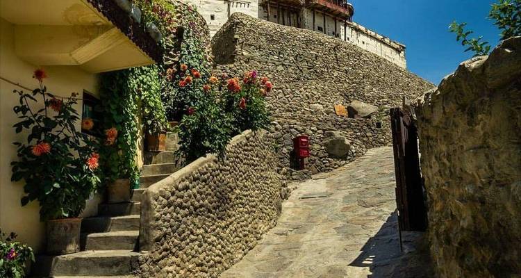 Callejón estrecho de piedra bordeado de flores en flor que conduce a un fuerte histórico bajo un cielo azul profundo.