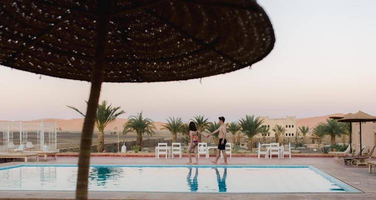 Two travelers walk beside a pool at a desert hotel with distant dunes and palms at twilight.