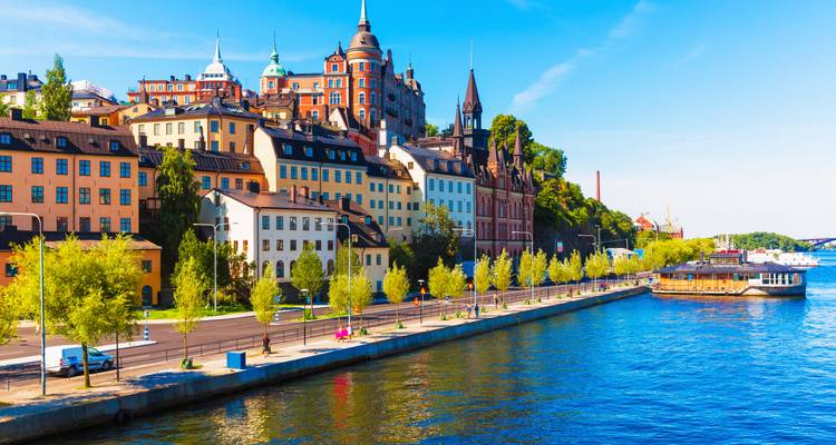 Vue pittoresque du front de mer présentant les bâtiments colorés de Södermalm et les arbres se reflétant sur l'eau bleue calme sous un ciel dégagé.