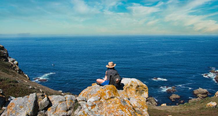 Traveller sitting on lichen-covered rocks gazing at the vast Atlantic Ocean along Ireland’s rugged coast.