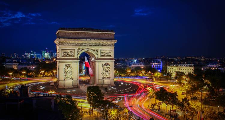 Arc de Triomphe nocturne avec des traînées de phares de voitures éclatantes circulant autour du monument.