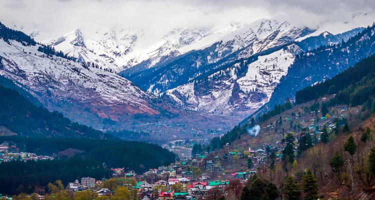 Snow-clad Himalayan peaks tower above a forested valley dotted with colorful village houses under moody clouds.