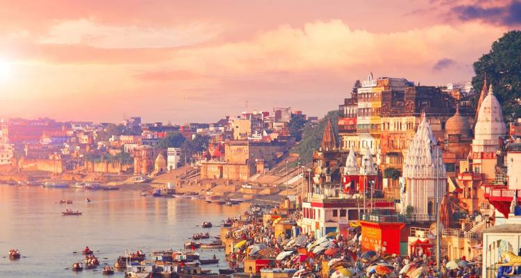 Panoramic view of Varanasi ghats along the Ganges at sunset with boats and colorful temples lining the riverbank.