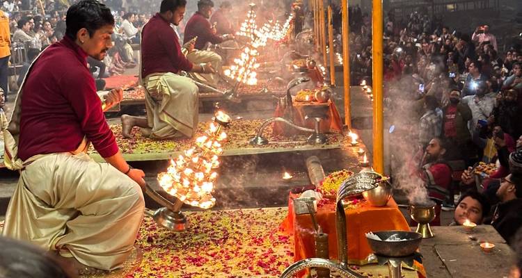 Priests on raised platforms wave flaming lamps while flower petals cover the stage at a crowded night aarti.