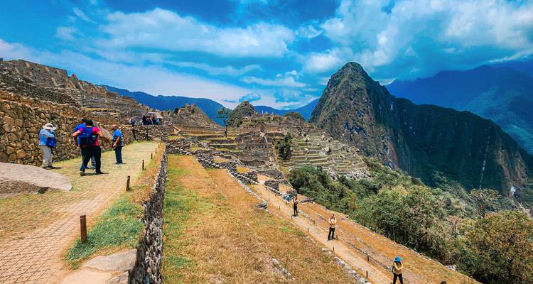 Bezoekers verkennen de terrassen van Machu Picchu met een indrukwekkende bergachtergrond onder een wolkige blauwe hemel.