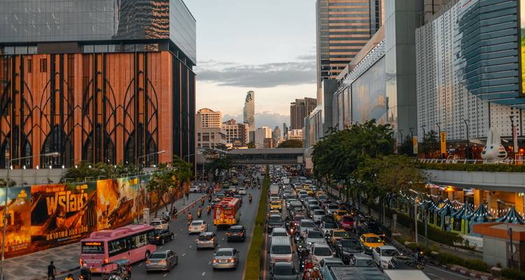 Dense traffic flows through a modern corridor of Bangkok skyscrapers at dusk.